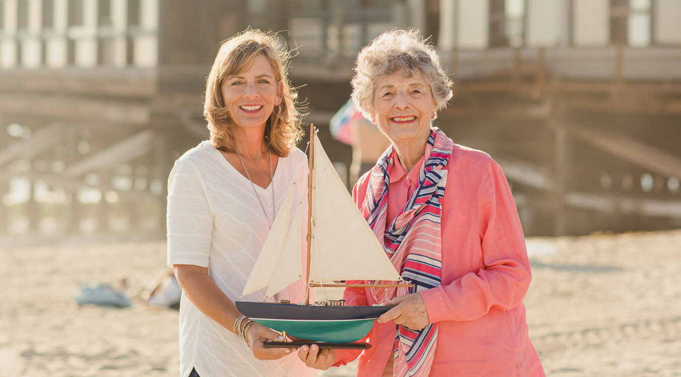 elderly mother and adult daughter on beach with sailboat smiling in the sunshine