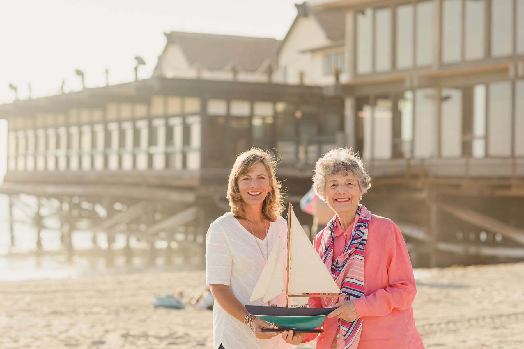 elderly mother and adult daughter on beach with sailboat smiling in the sunshine