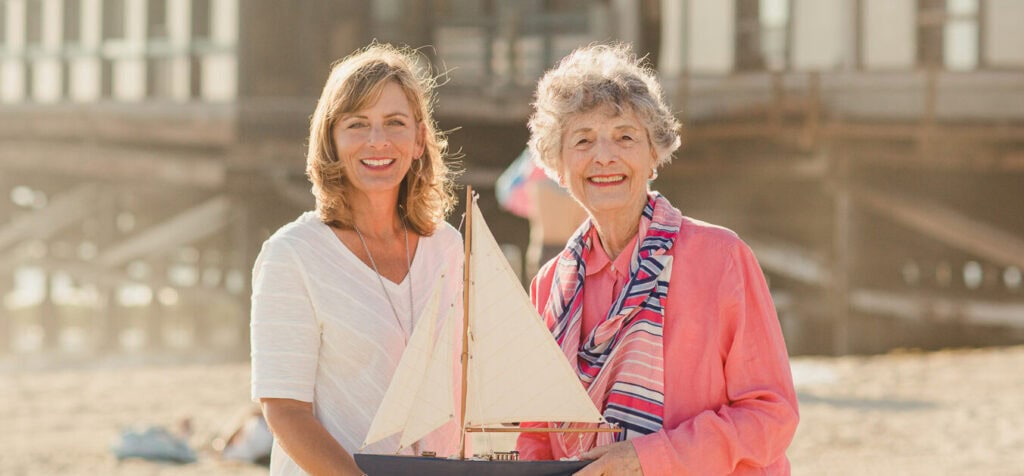 elderly mother and adult daughter on beach with sailboat smiling in the sunshine